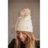 Penelope Michka cable knit pom-pom hat with wide cuff and diamond-shaped logo plate.